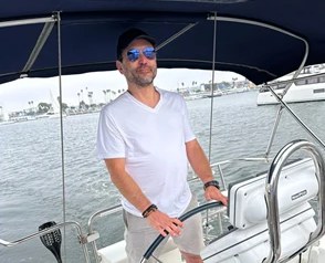 Yener Yardim standing at the wheel of his sailboat in a marina, in a white T-shirt and a cap.