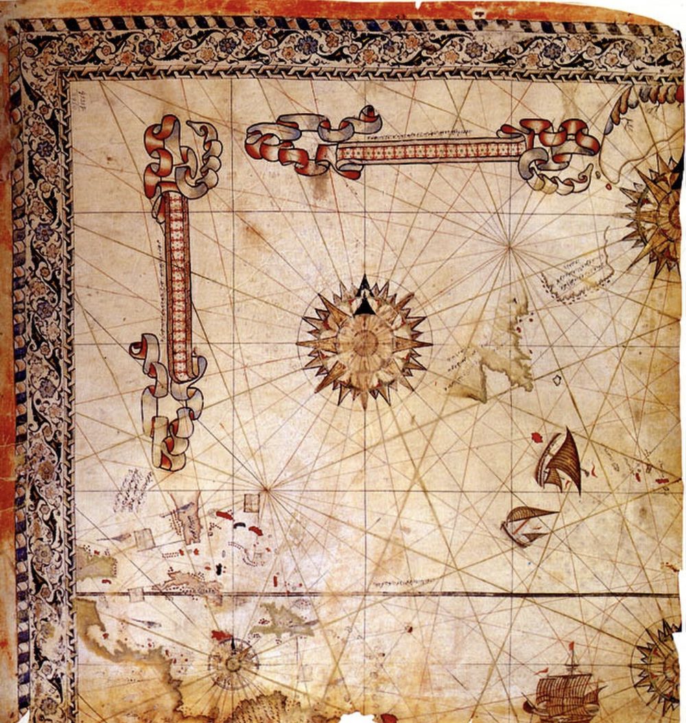 A fragment of Piri Reis's 1528 world map on parchment: a decorated floral border, large compass roses with rhumb lines, ships under sail, and the islands of the Caribbean at the lower left.